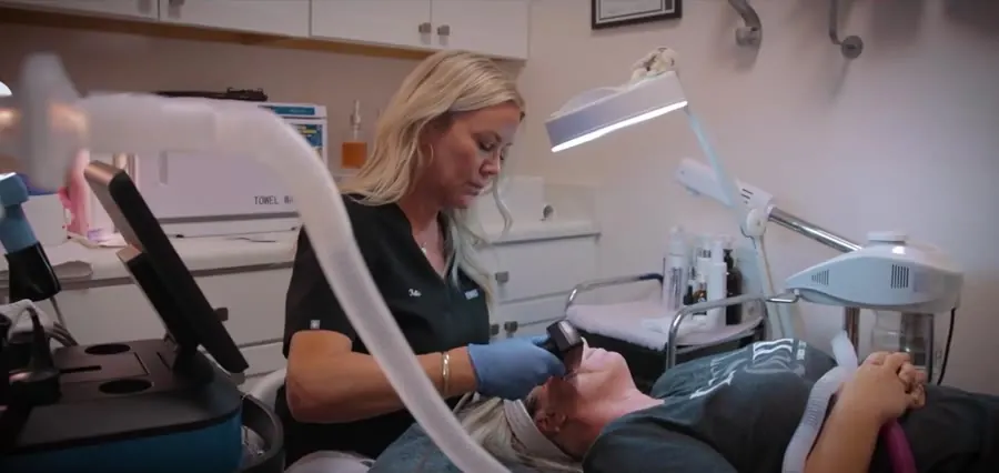 A provider guiding a laser handpiece over a patient reclined under a treatment lamp